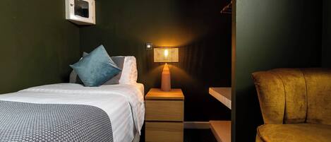A hotel room with a bed, a nightstand with a lamp, a chair, and a small cabinet with a picture.