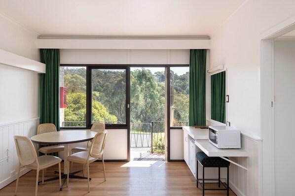 A modern kitchen with a dining area, large windows with green curtains, and a view of a forested area.