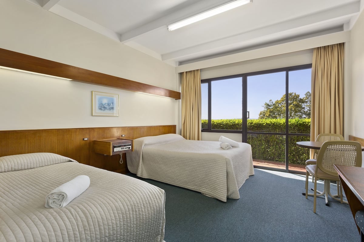 A hotel room with two beds, a desk, a chair, and a large window with a view of greenery.