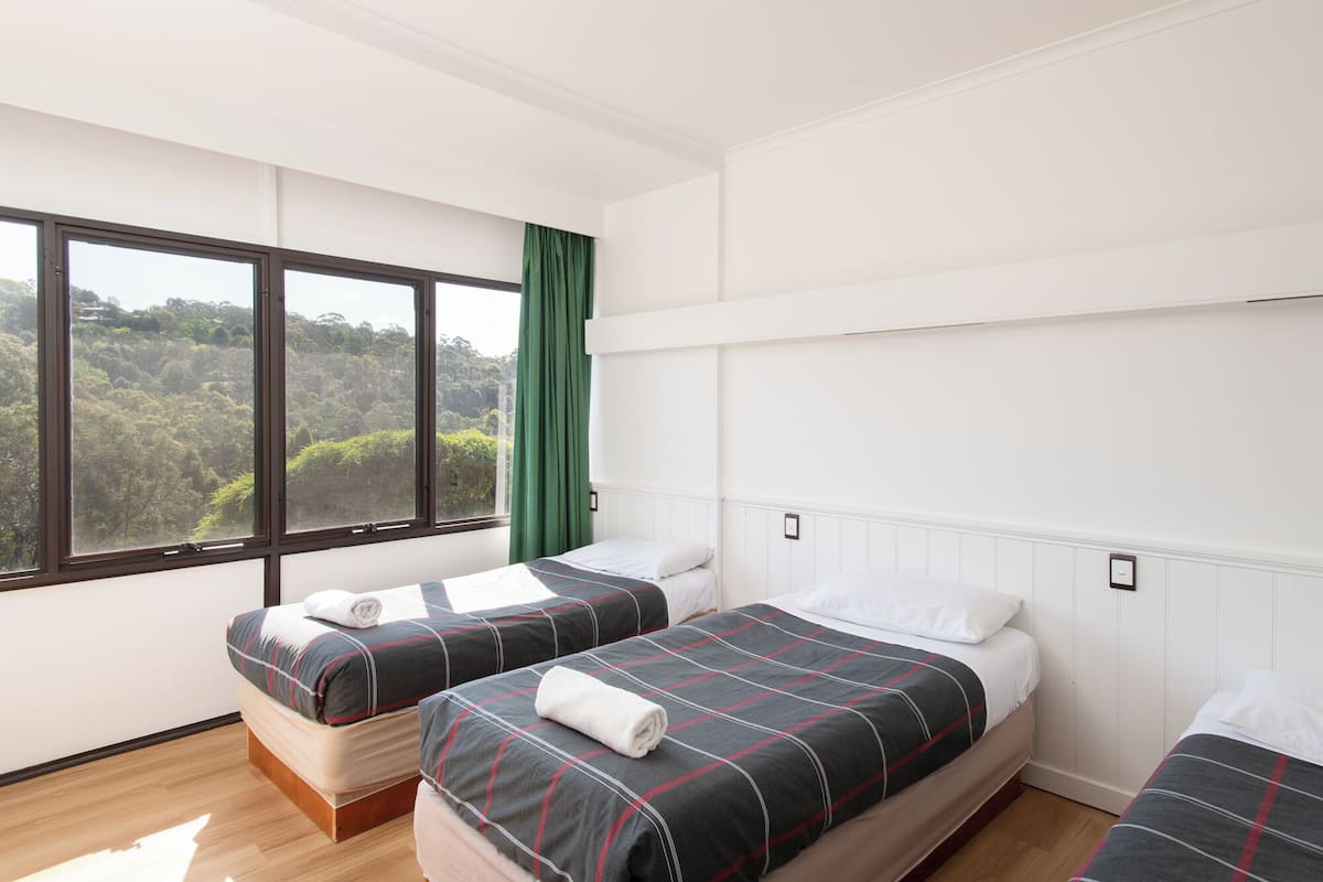A hotel room with two beds, a large window with a view of a forest, and a green curtain.