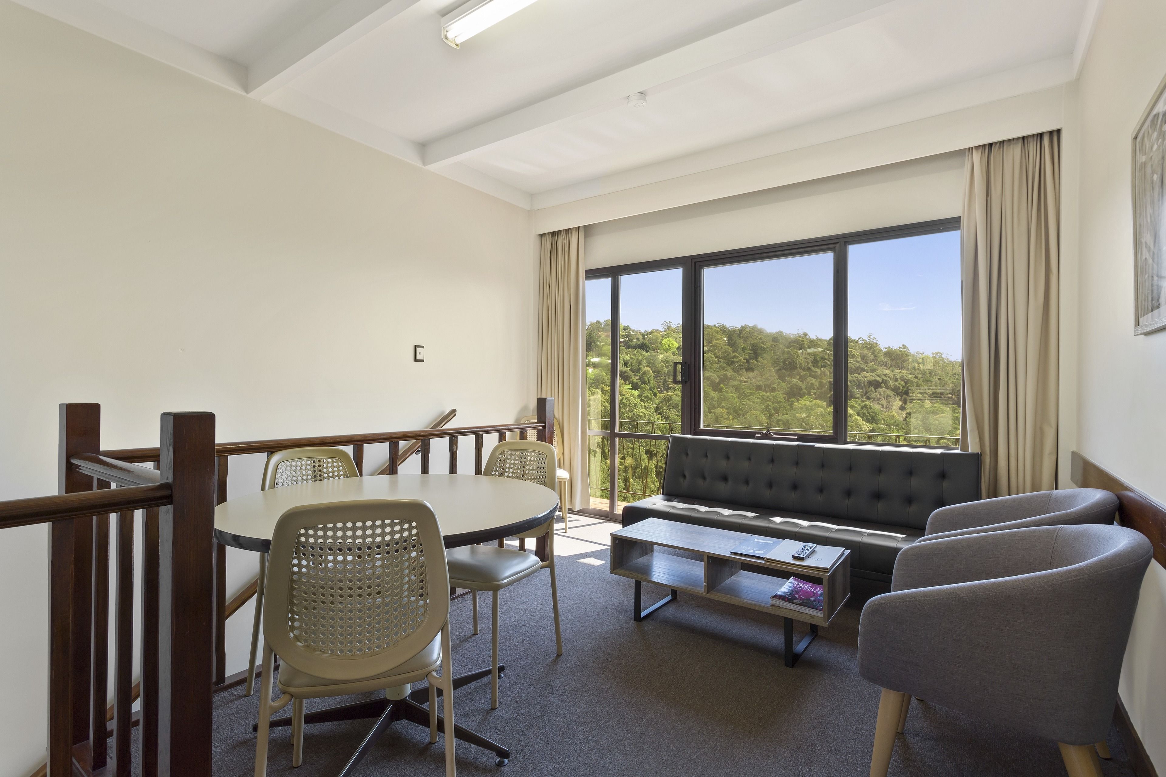 A modern room with a dining table, chairs, a sofa, and a large window offering a view of greenery.