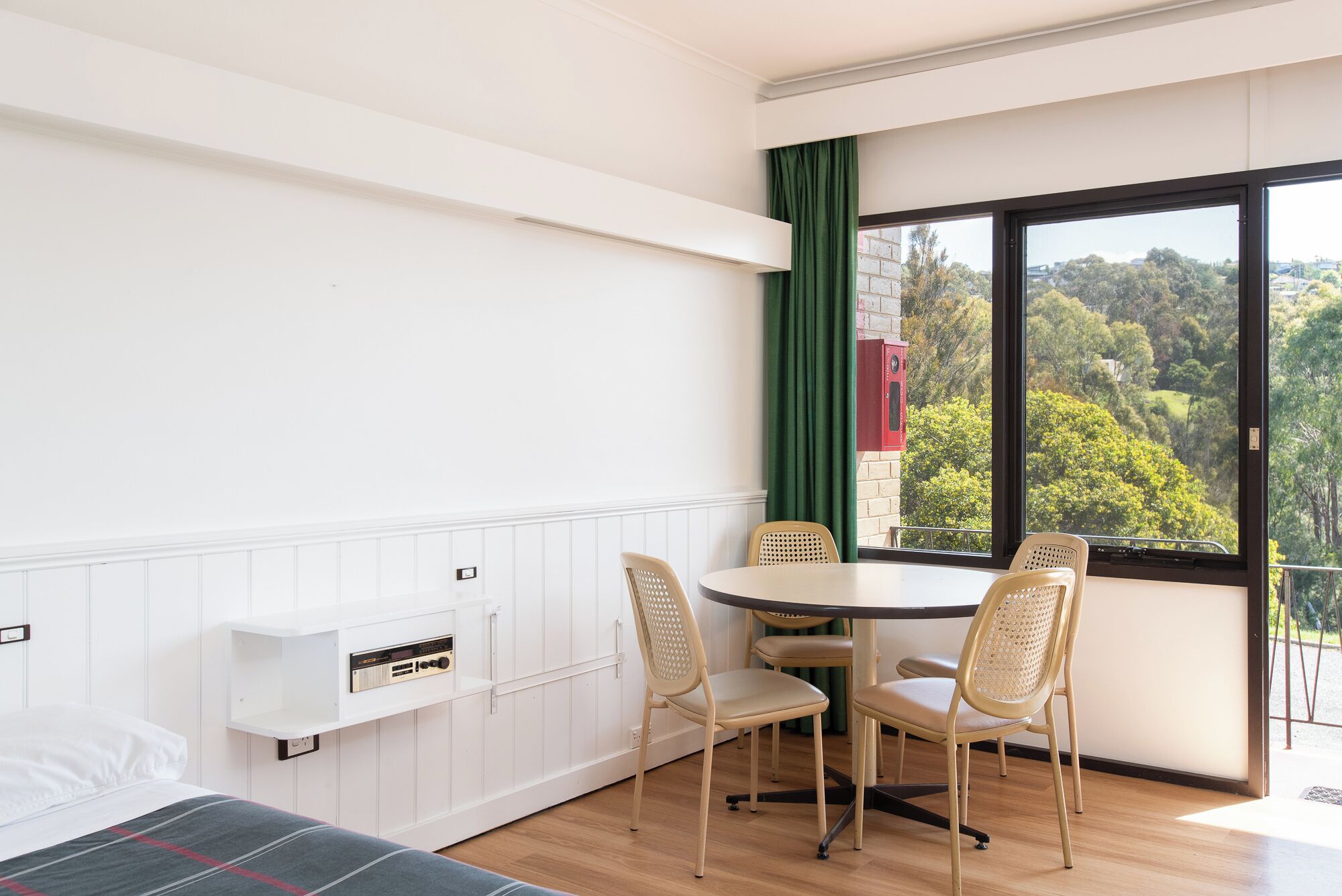 Une pièce comprenant un lit, une table ronde avec quatre chaises, une radio et une vue sur un paysage verdoyant par le biais d’une grande fenêtre.