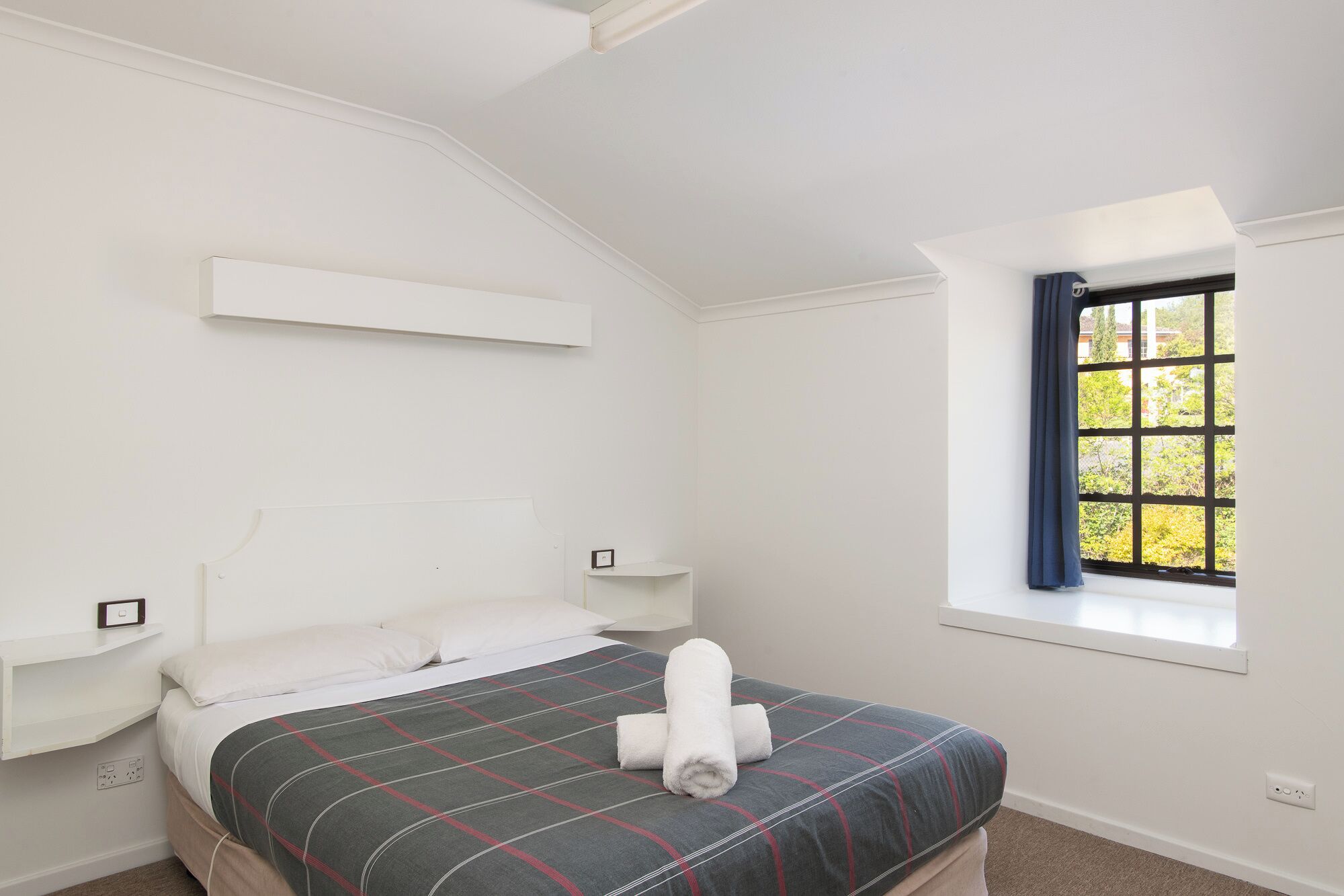 A bedroom with a bed, a window with curtains, a bedside table, and a wall-mounted shelf.