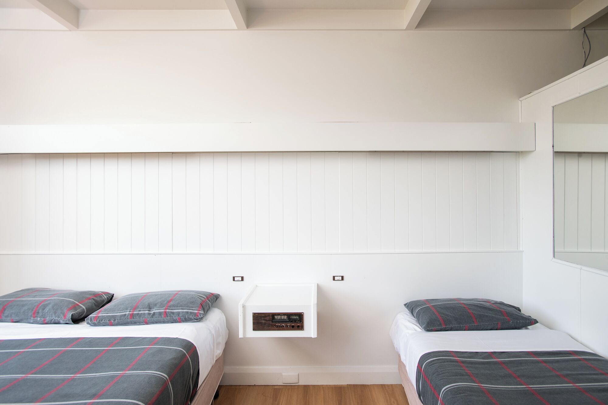 A bedroom with two beds, each with a grey and red plaid bedspread, a white wall, and a small white shelf with a clock.
