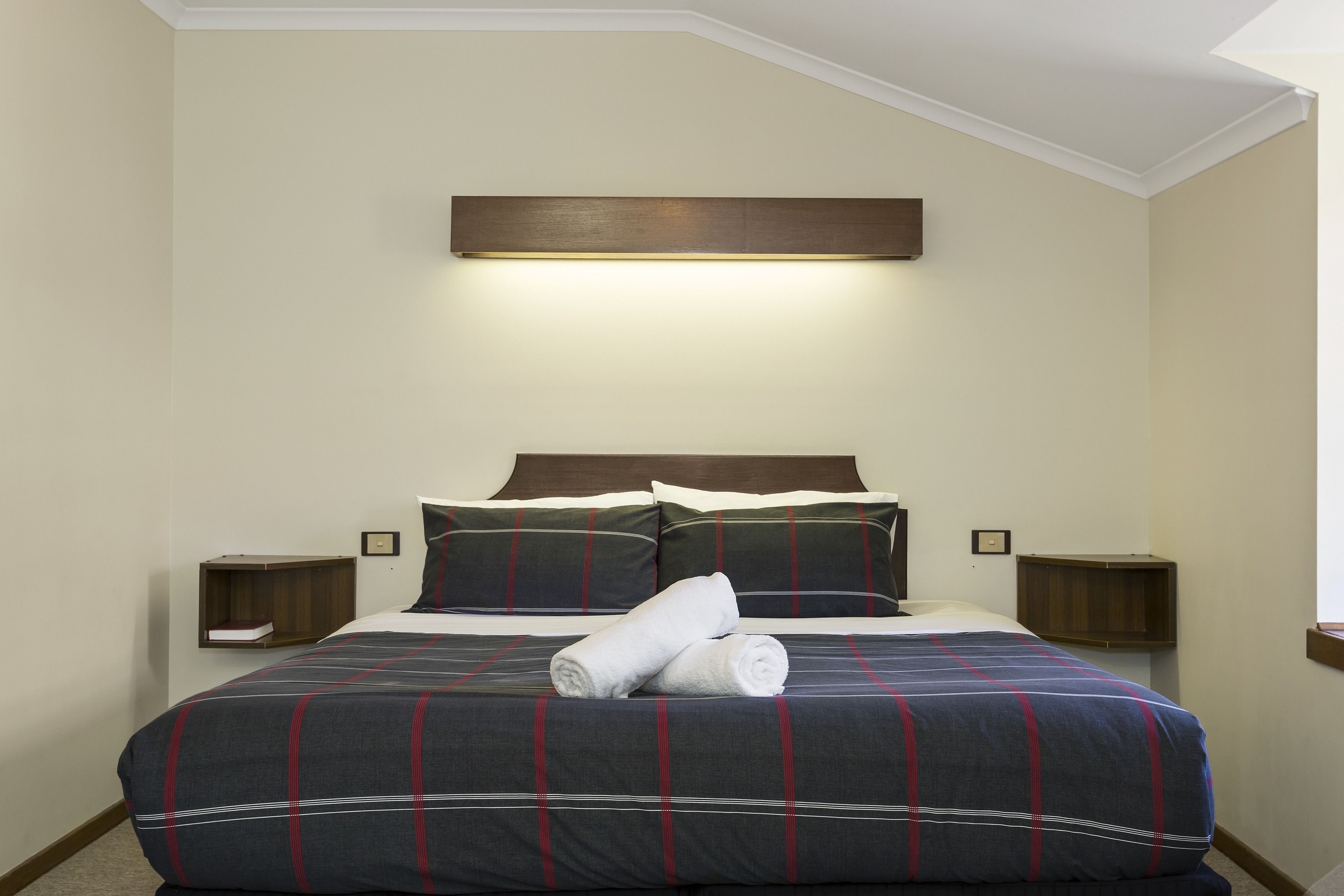 A neatly made bed with a dark blue and red striped bedspread, two pillows, and a rolled towel on the bed. There are two bedside tables with lamps on either side of the bed.