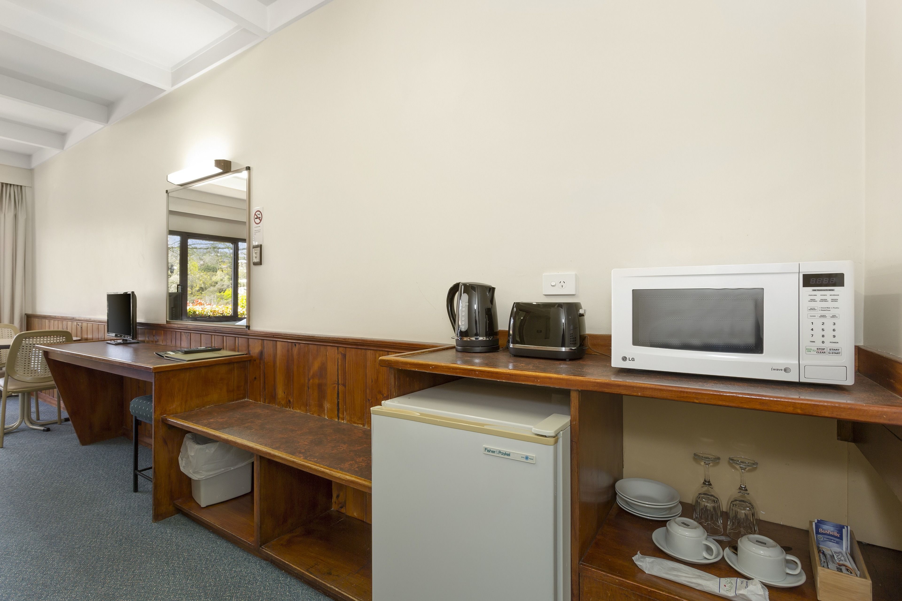 A hotel room with a wooden desk, a refrigerator, a microwave, a coffee maker, and a window with a view of trees.