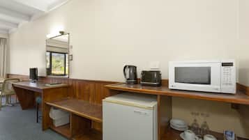 A hotel room with a wooden desk, a refrigerator, a microwave, a coffee maker, and a window with a view of trees.