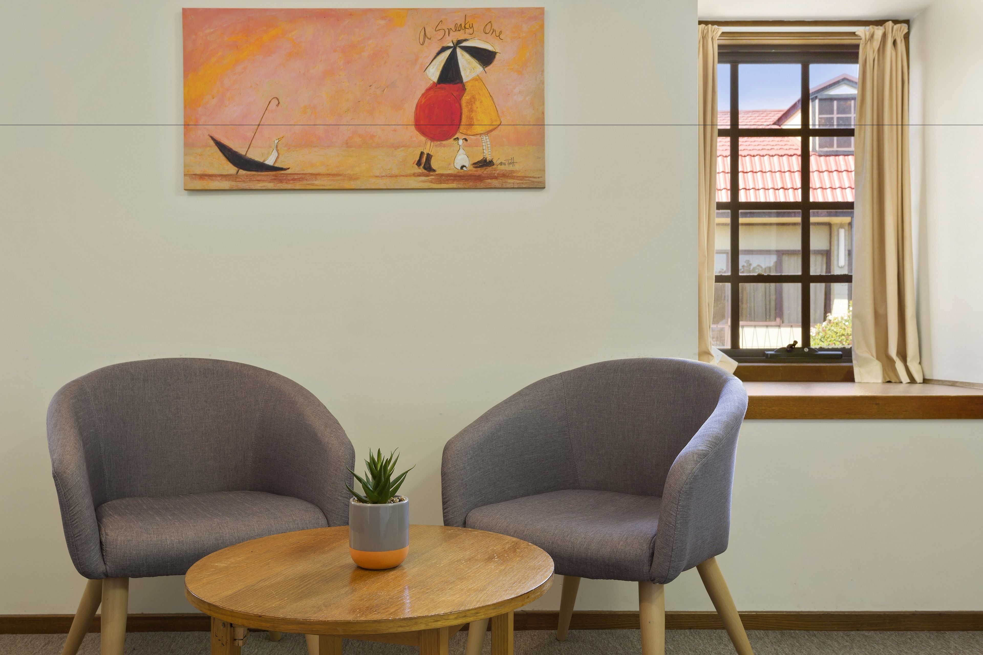 A room with two grey armchairs, a wooden table with a potted plant, and a painting above the window.