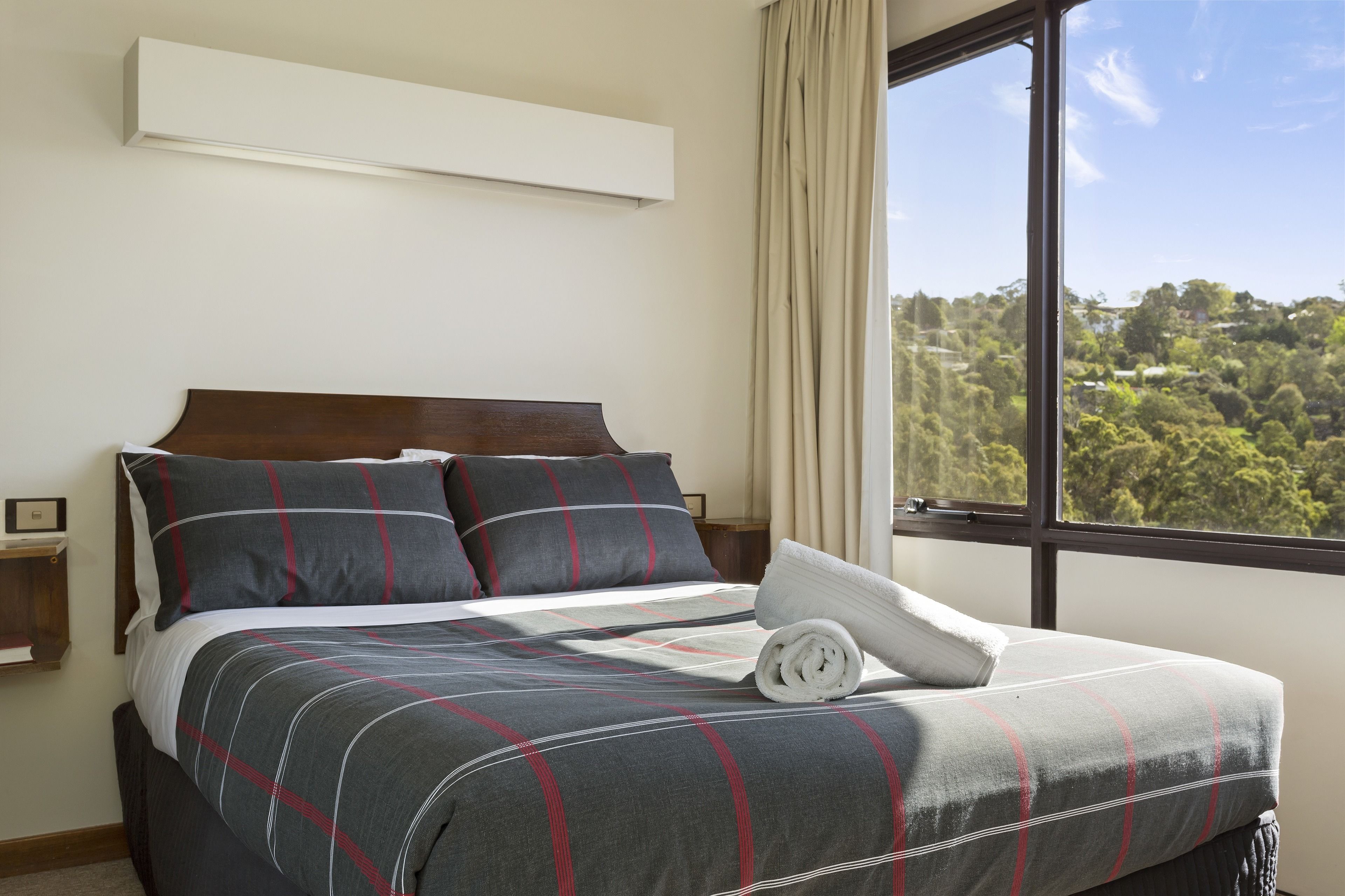 Una cama bien tendida con una colcha gris y roja a cuadros, un cabecero de madera y vistas a un paisaje verde a través de un ventanal grande.