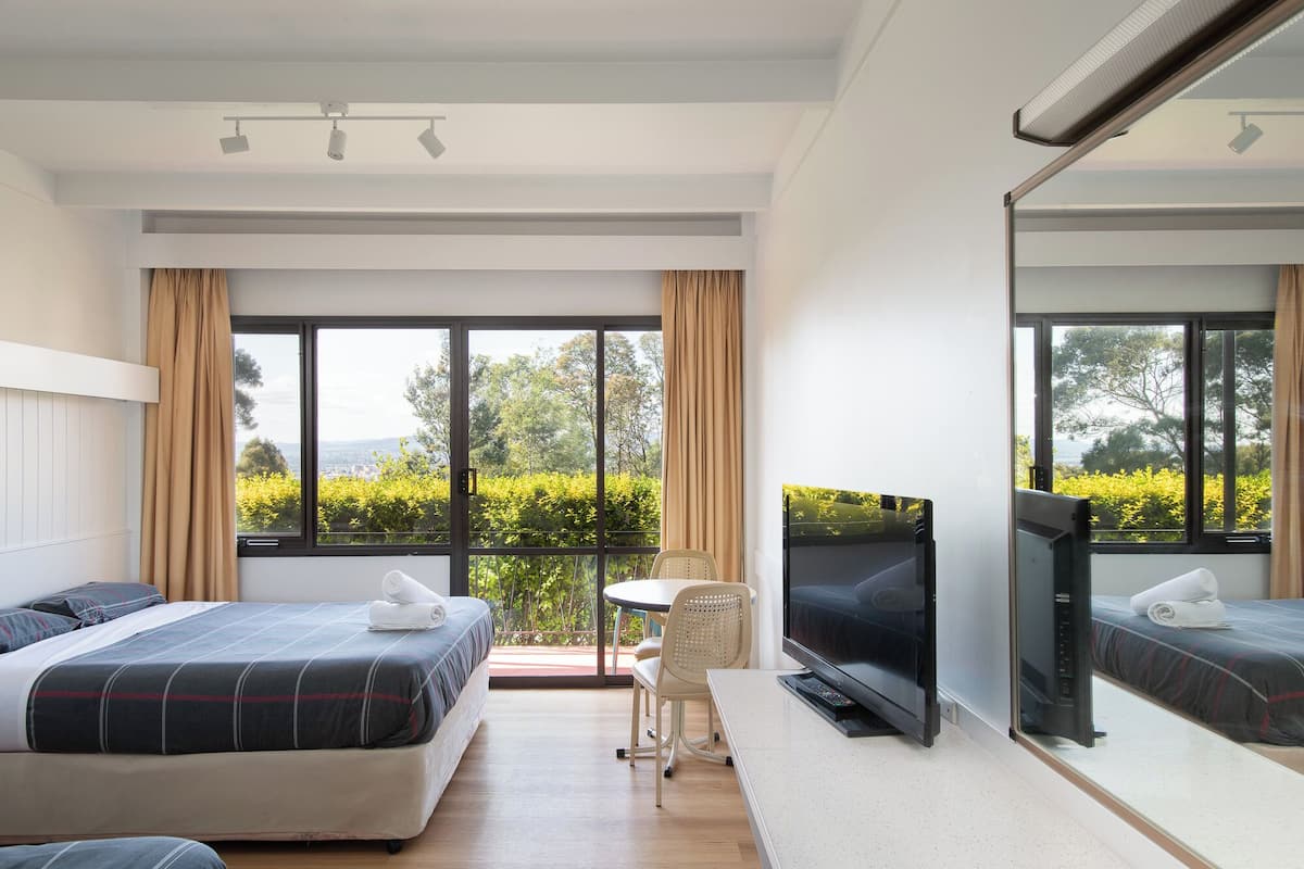 A modern hotel room with a large bed, a TV, a small table, and a balcony with a view of greenery.