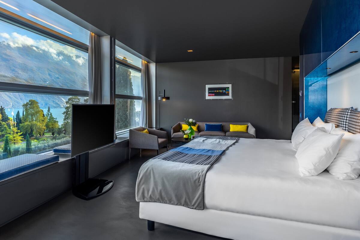 A modern hotel room with a large bed, a flat-screen TV, a sofa, and a view of a lake.