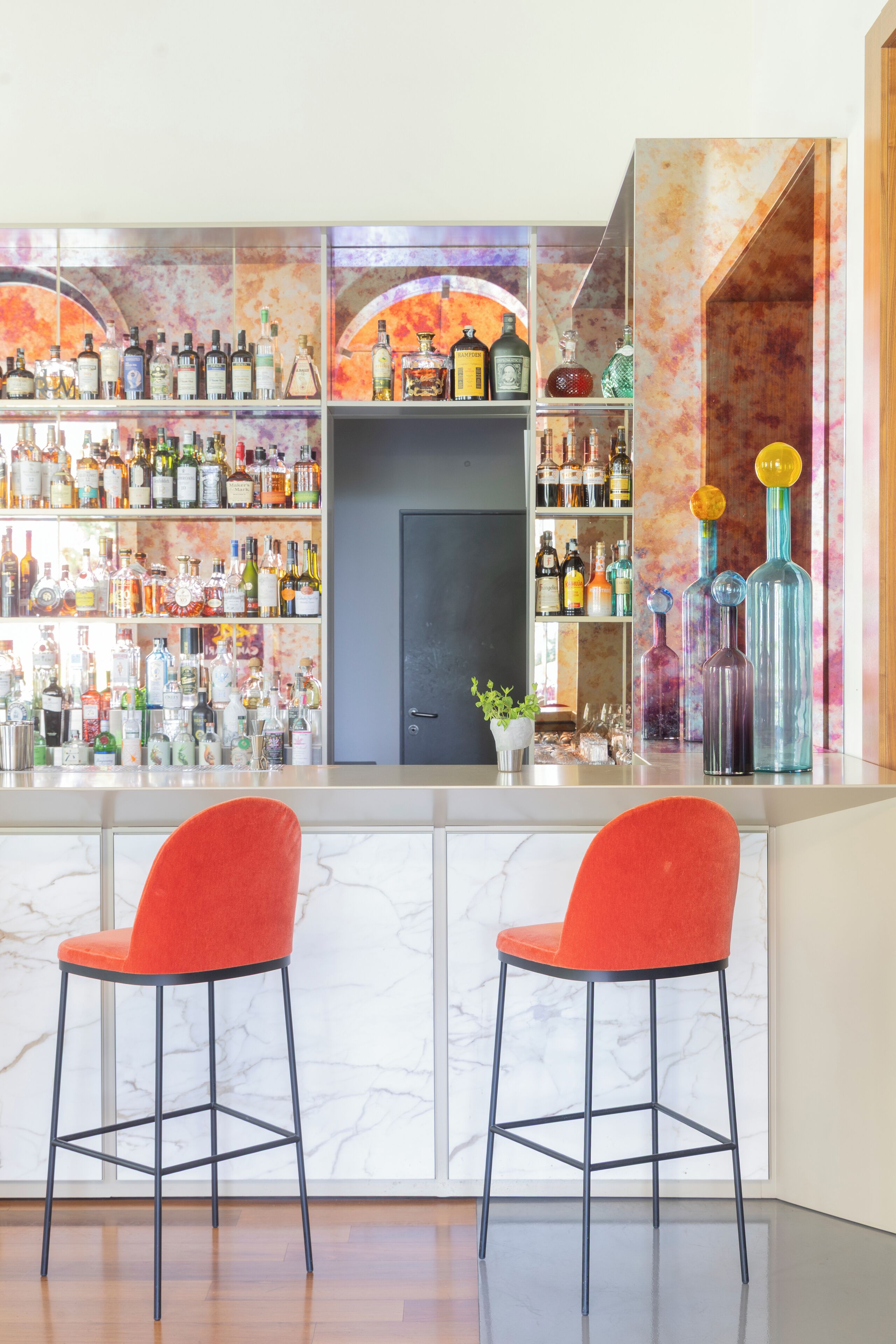 A bar area with two red bar stools, a marble wall, and shelves stocked with various liquor bottles.