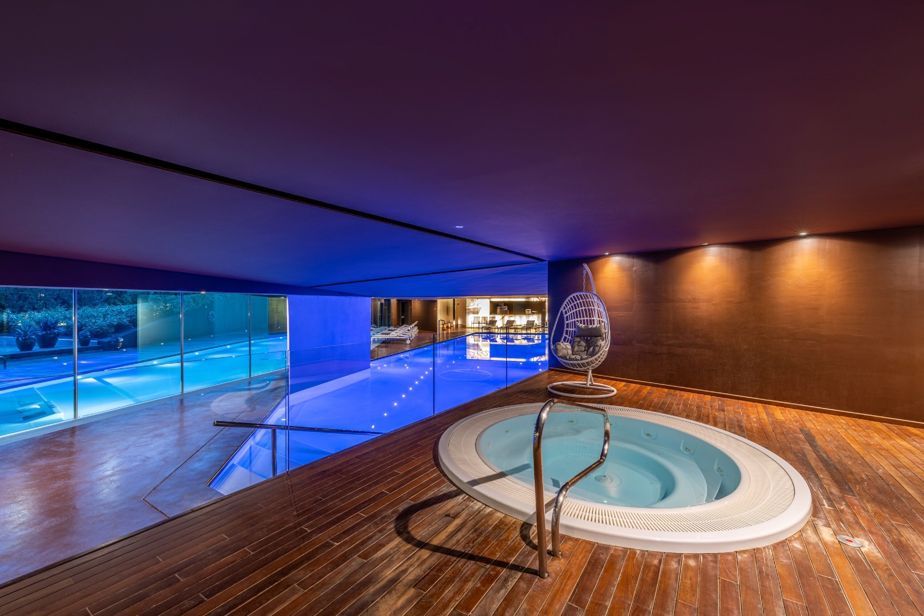 Un'area piscina moderna in interni, con pavimento in legno, vasca idromassaggio e parete vetrata con vista sull'esterno.