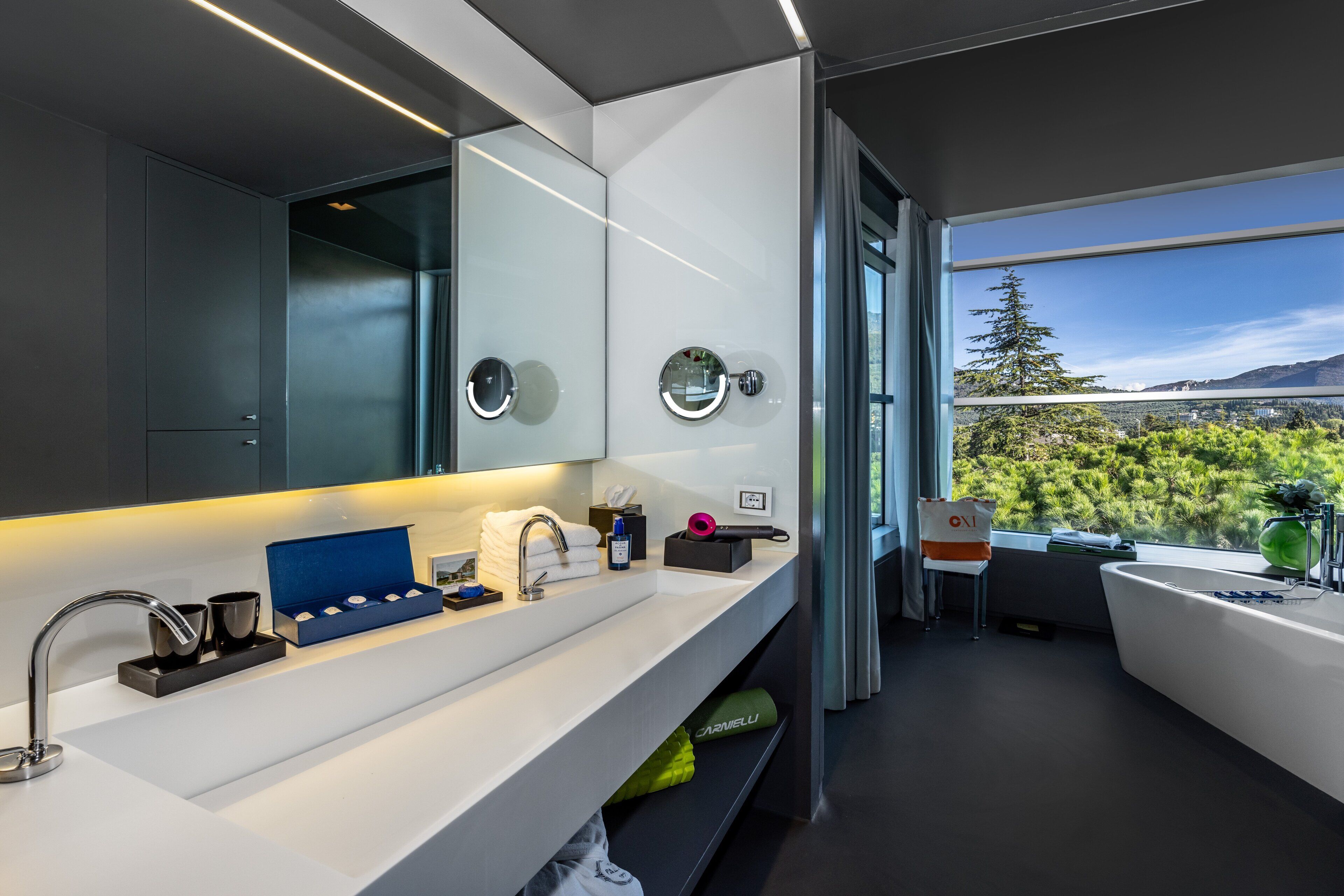 Une salle de bain moderne avec un grand miroir, une baignoire et une vue sur une forêt, visible à travers une grande fenêtre.
