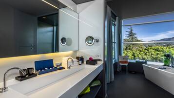 Une salle de bain moderne avec un grand miroir, une baignoire et une vue sur une forêt, visible à travers une grande fenêtre.