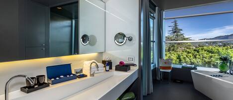 Un baño moderno con un espejo grande, una bañera y vistas a un bosque a través de un ventanal amplio.