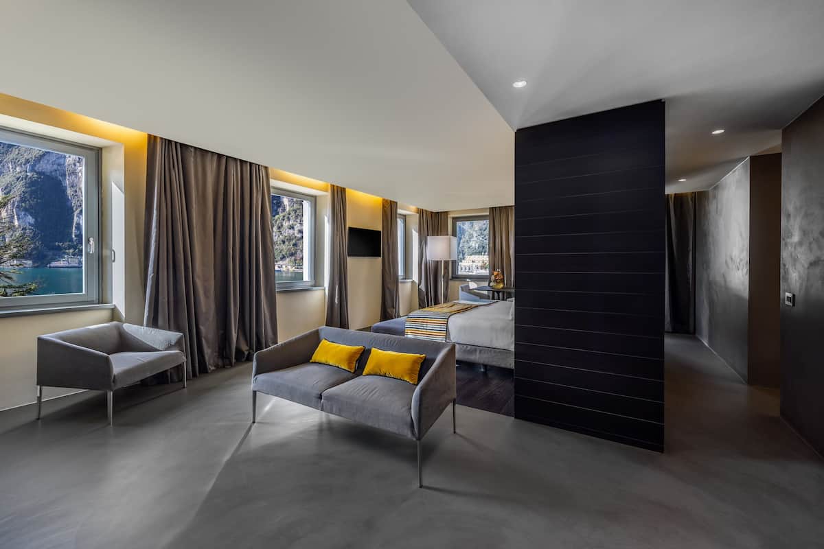 A modern hotel room with a large bed, a sofa with yellow cushions, and a view of the sea through large windows.