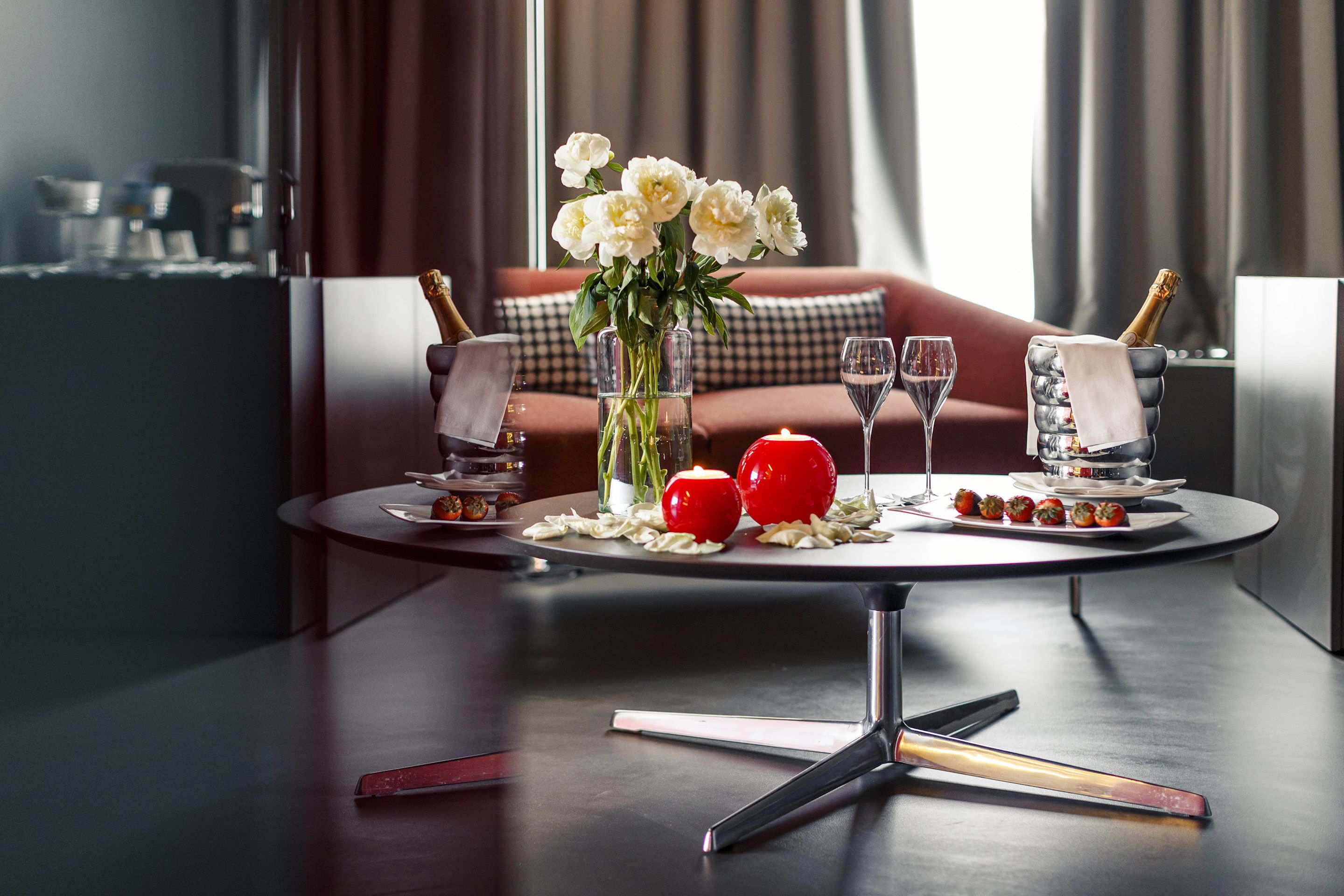 A modern dining table with a vase of flowers, two red apples, a bottle of champagne, and a glass of wine.