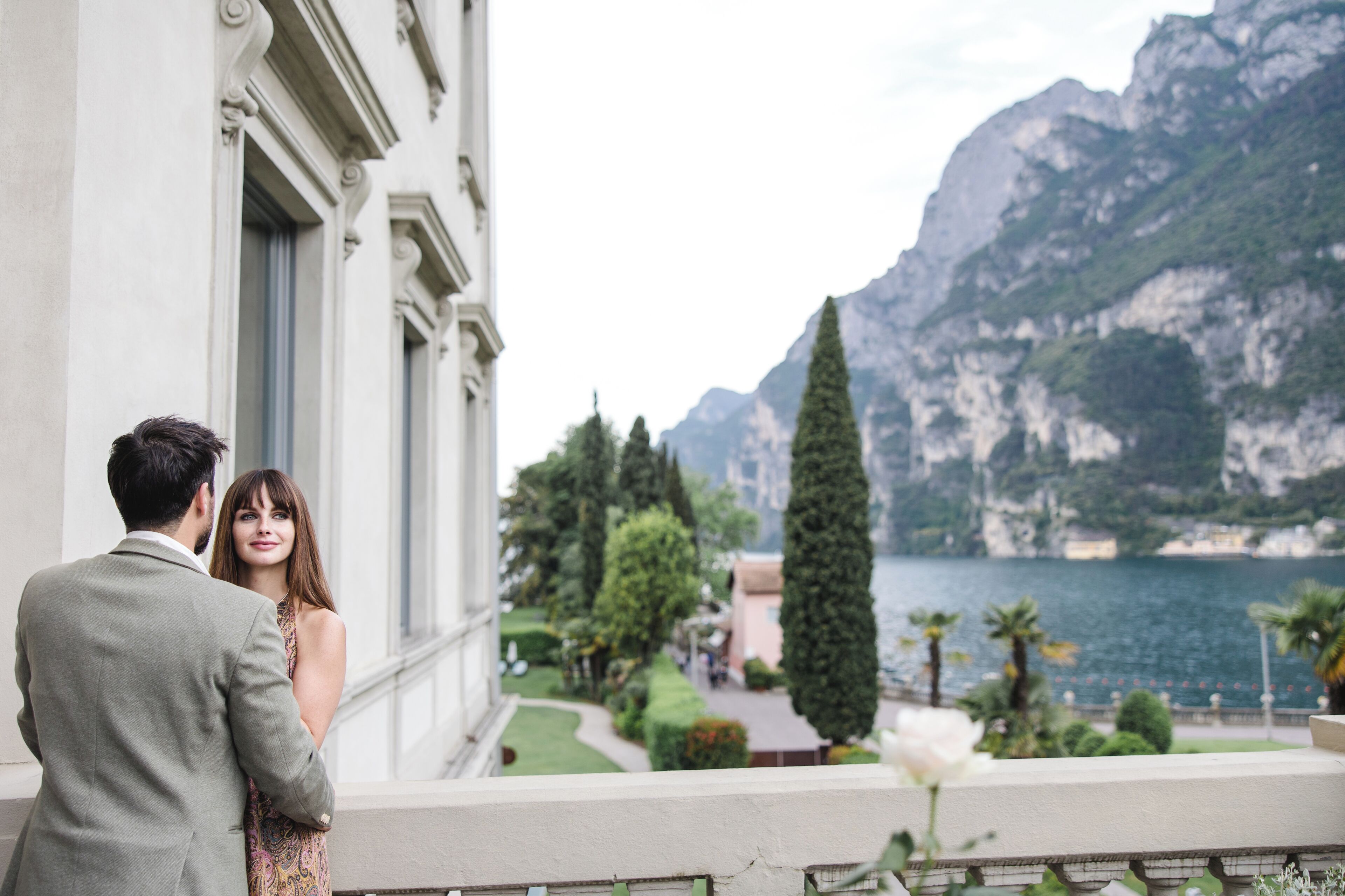 A couple standing by a balcony overlooking a mountainous lake with a garden and a white building in the background.