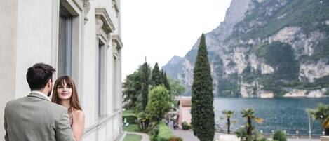 A couple standing by a balcony overlooking a mountainous lake with a garden and a white building in the background.