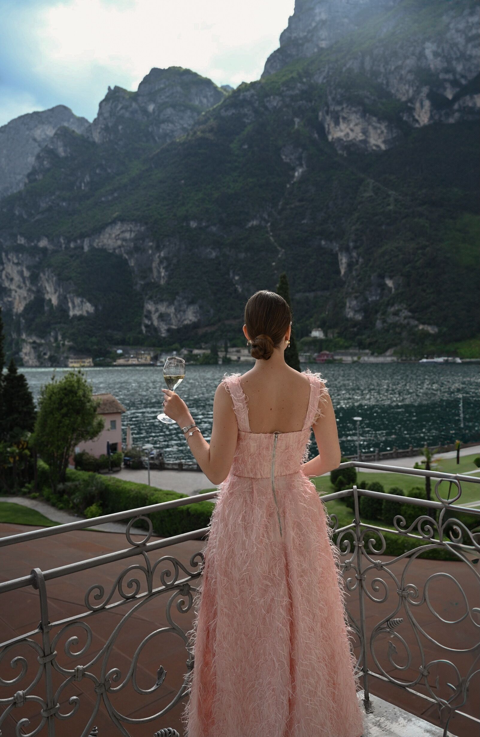 Una mujer con un vestido rosa de pie en un balcón con vista a un lago y montañas.