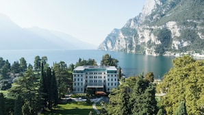 A large, white building with a symmetrical facade, surrounded by lush greenery and a mountainous backdrop. - Lido Palace - The Leading Hotels of the World (Riva del Garda)