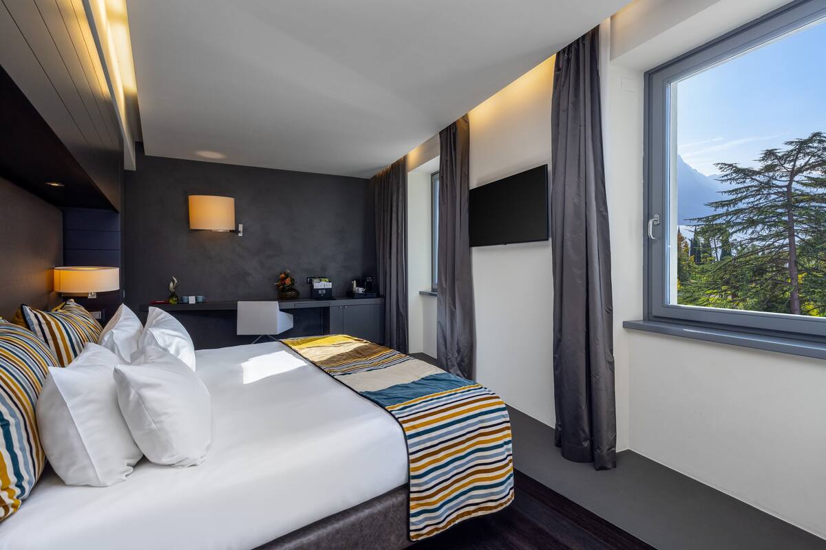 A modern hotel room with a large bed, a bedside table, a flat-screen TV, a window with curtains, and a view of trees and mountains.