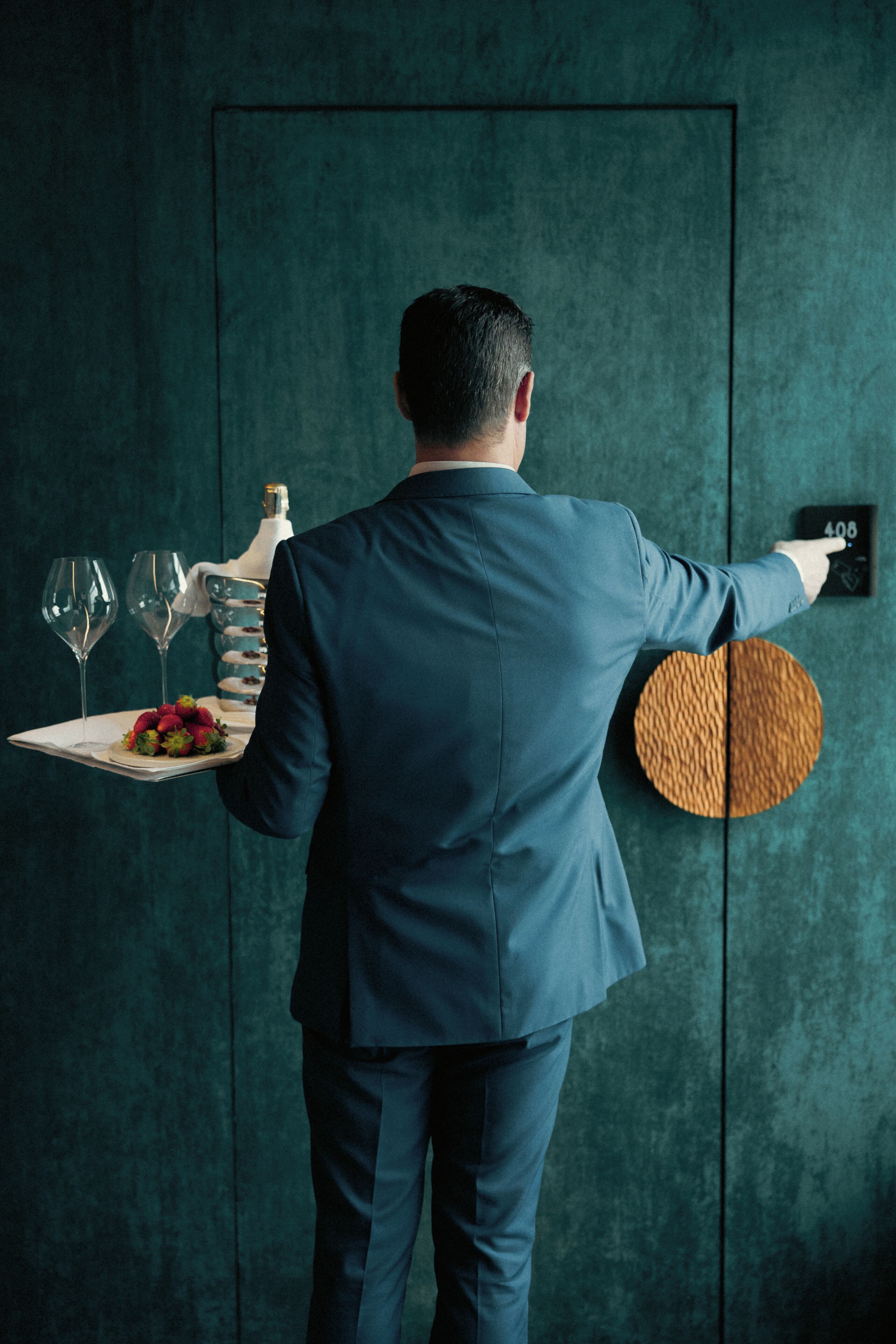 A person in a blue suit holding a tray with a bottle, glasses, and a plate of food.