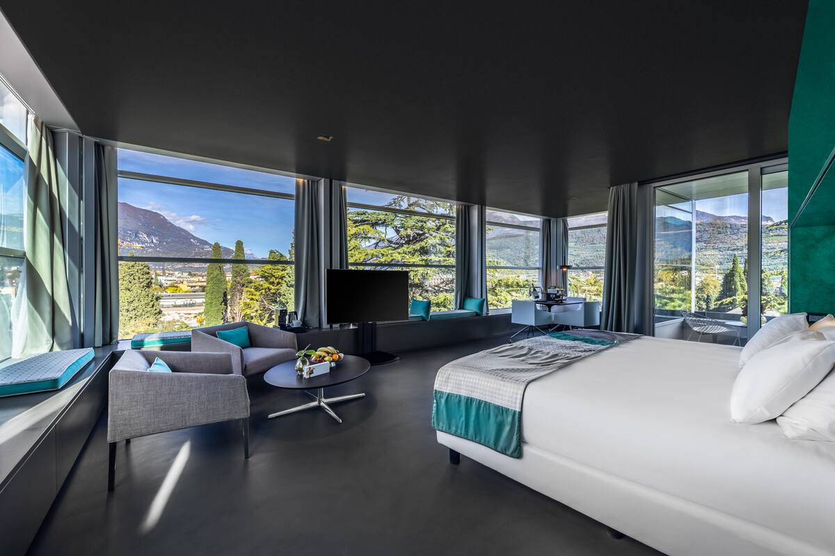 A modern hotel room with a large bed, a sofa, a desk, and a TV. The room has large windows offering a mountain view.