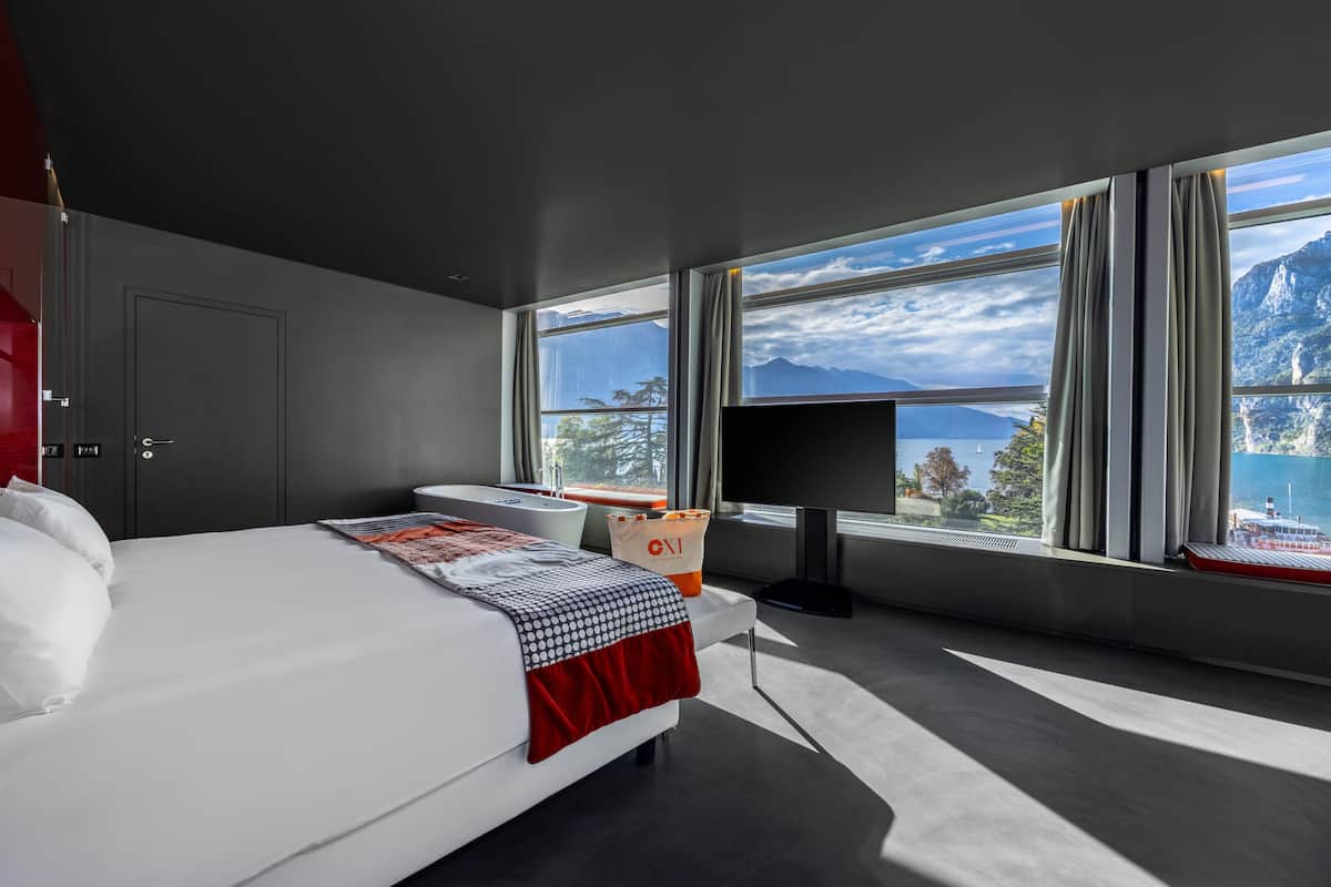 A modern hotel room with a large bed, a TV, and a view of mountains and a lake.