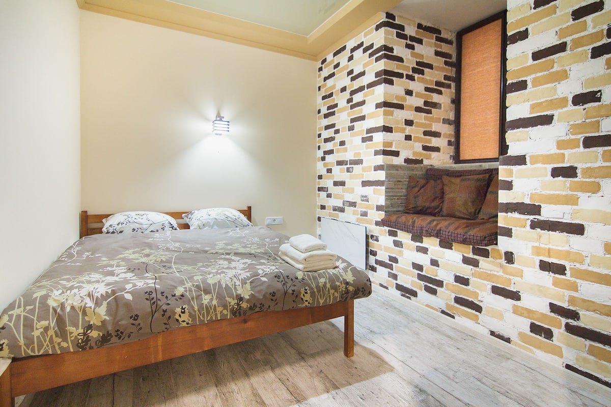 A bedroom with a wooden bed, a brick-patterned wall, and a small seating area with a cushion.
