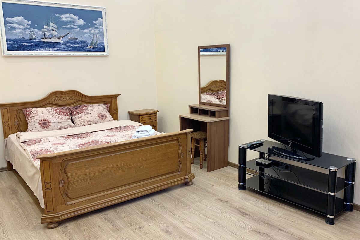 A bedroom with a wooden bed, a television on a stand, a nightstand, a mirror, and a painting of a sailboat on the wall.