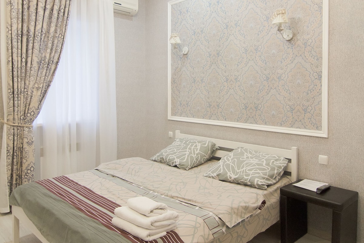 A bedroom with a bed, a nightstand, a mirror, a chandelier, and a window with curtains.
