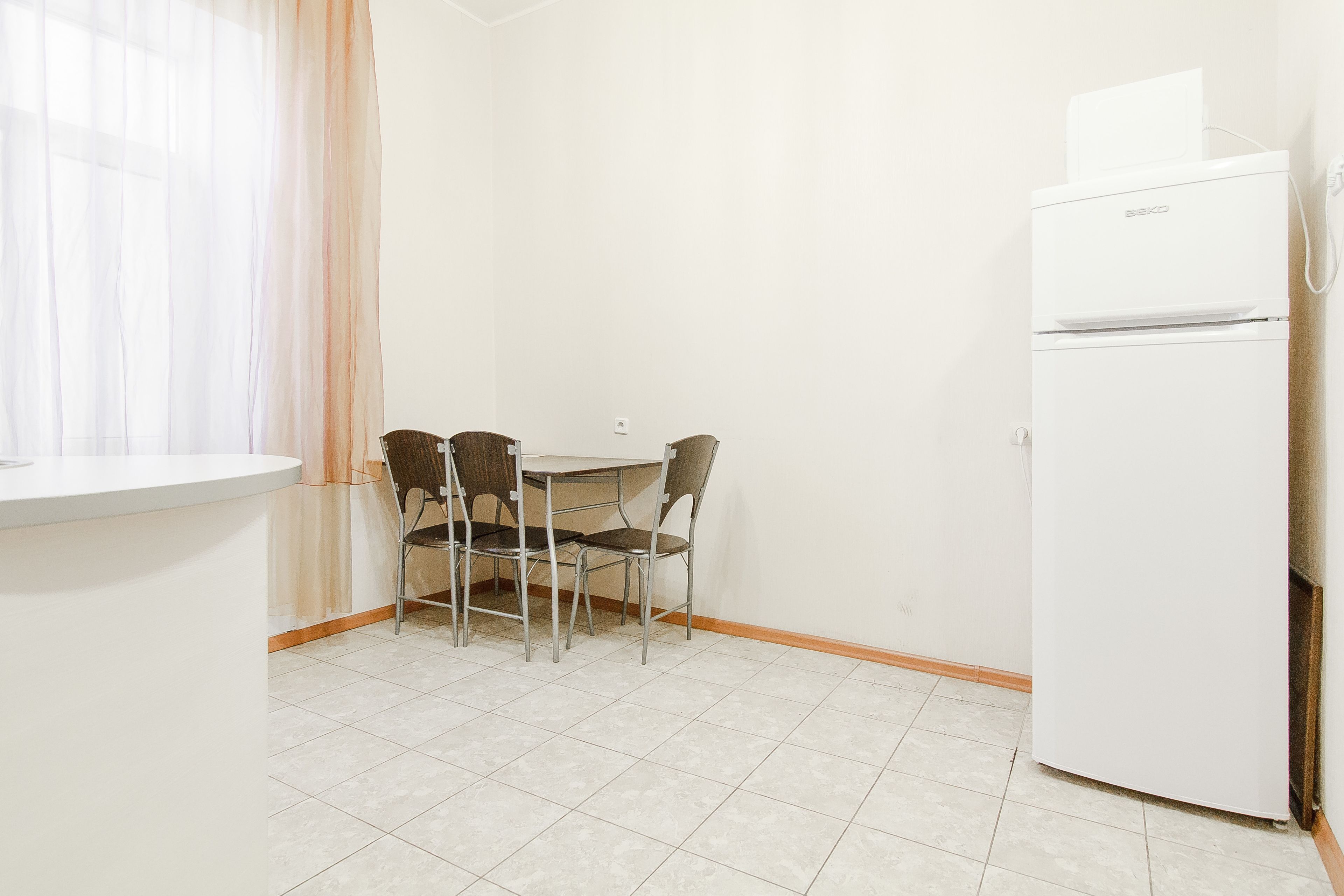 A small kitchenette with a white refrigerator, a table with four chairs, and a window with sheer curtains.