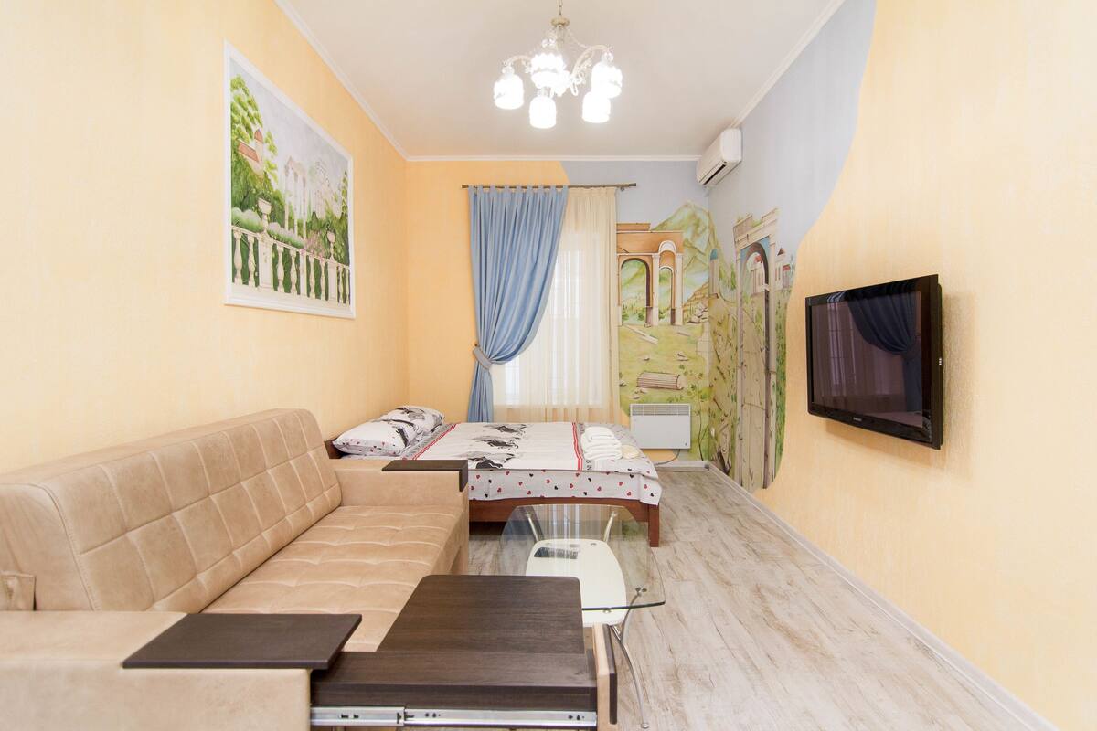 A small, cozy room with a sofa, a bed, a TV, and a desk with a laptop.