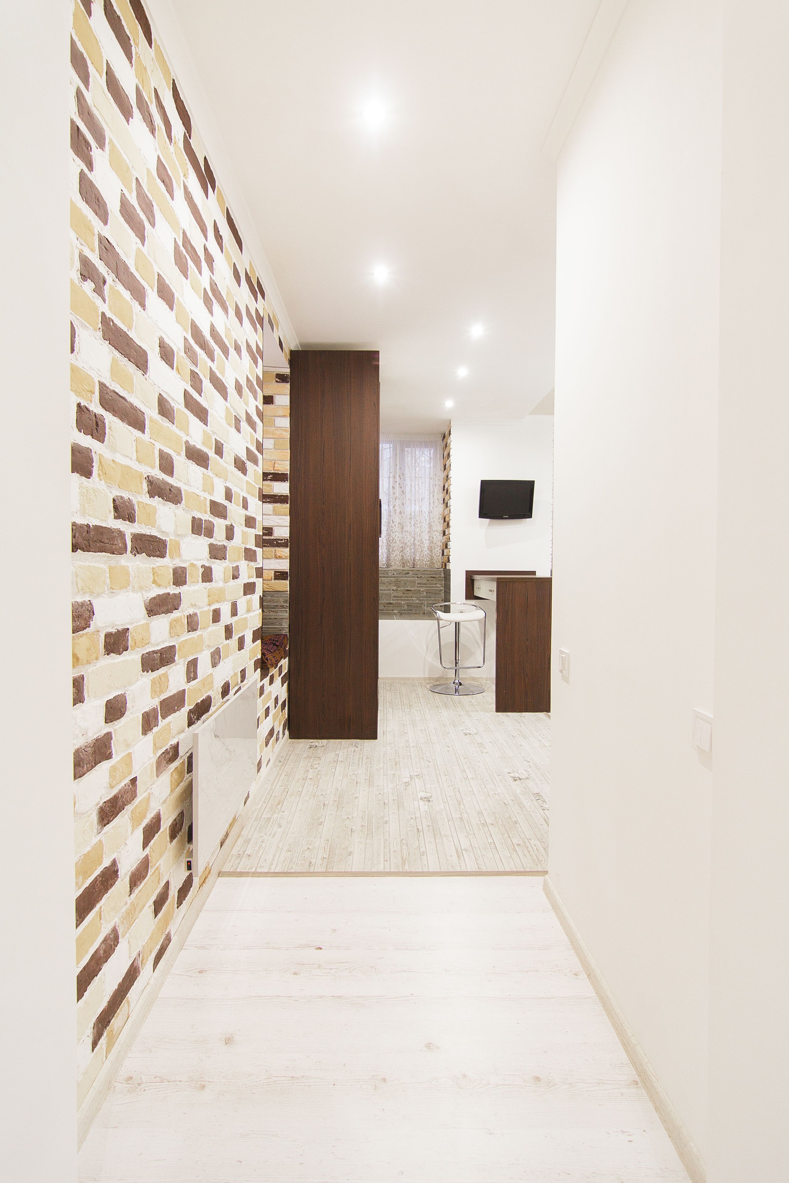 A hallway with a brick-patterned wall, a wooden door, a small round table, and a flat-screen TV mounted on the wall.