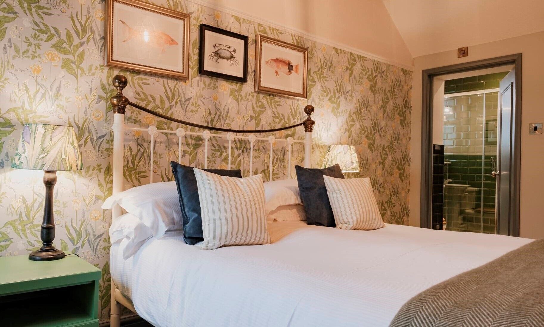 A hotel room with a bed, two nightstands, a green nightstand, a wall with floral wallpaper, and framed pictures.