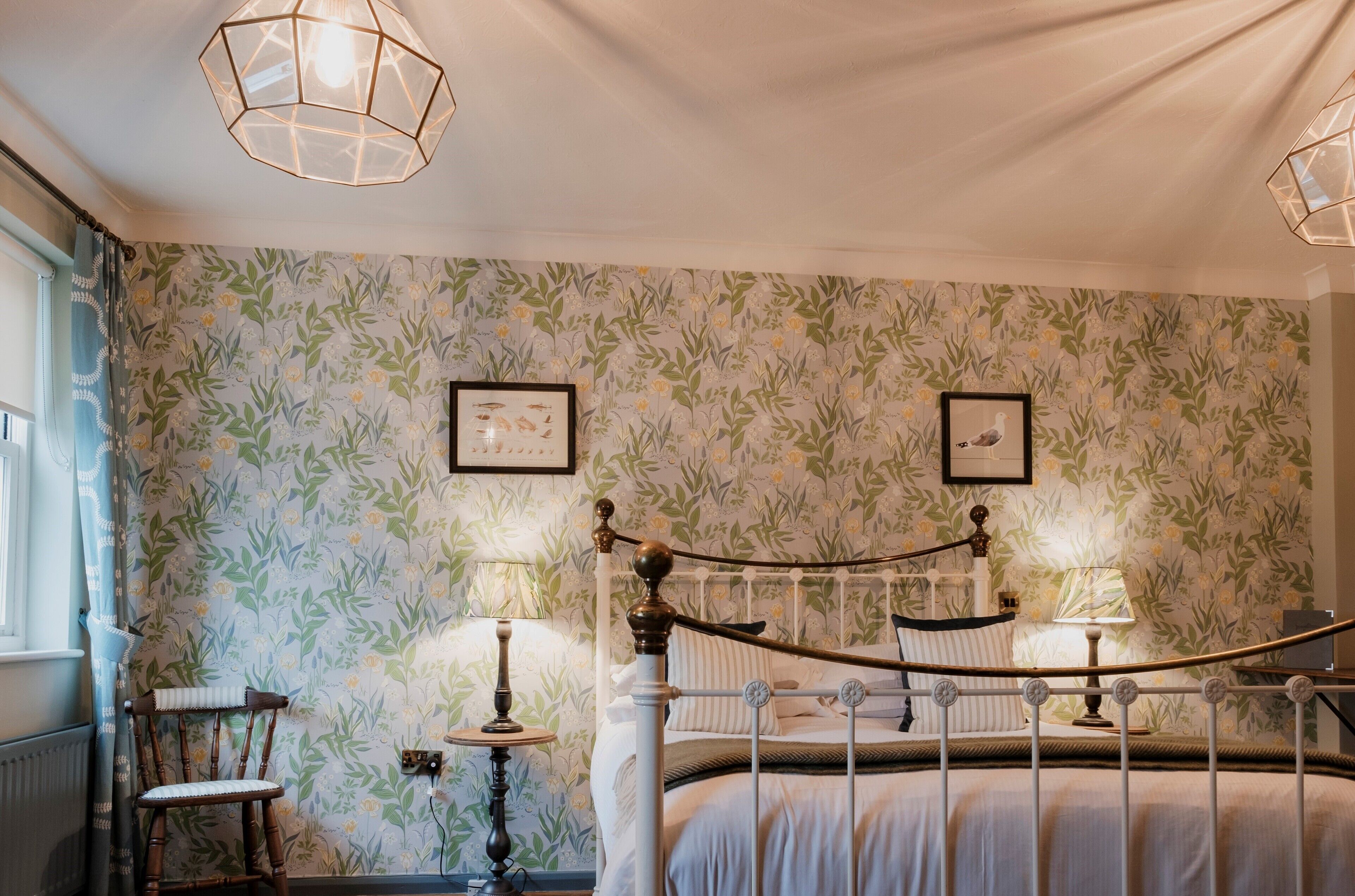 Un dormitorio con papel tapiz floral, una cama con estructura metálica, dos lámparas de noche y una silla de madera.