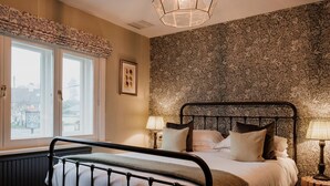 A bedroom with a bed, two bedside lamps, a window with a view, and a patterned wallpaper. - The Fisherman's Haunt (Christchurch)