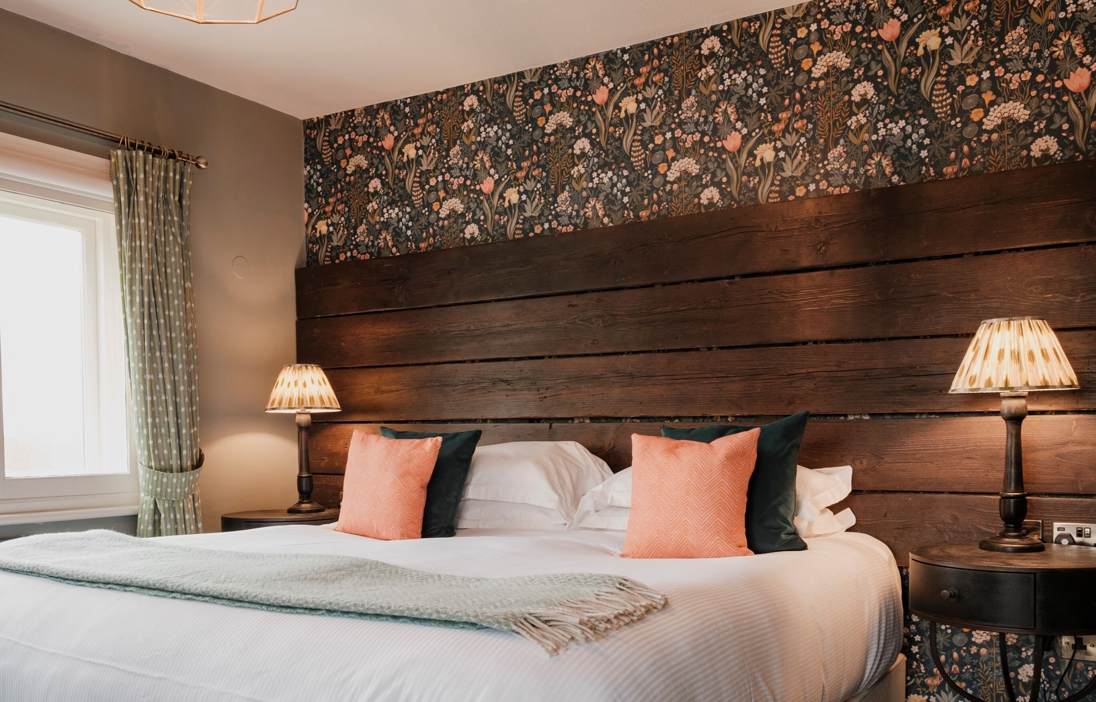 Ein Schlafzimmer mit einem großen Bett, zwei Nachttischen, einem Holz-Kopfteil, blumigem Wandpapier und einem Fenster mit Vorhängen.