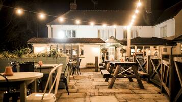 An outdoor patio area with stone flooring, wooden tables and chairs, and string lights.