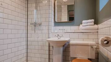 Un baño con azulejos blancos tipo metro, ducha con mampara de vidrio, lavamanos blanco, inodoro con asiento de madera y espejo con marco dorado.
