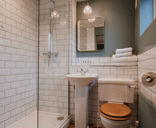 A bathroom with white subway tiles, a glass-enclosed shower, a white sink, a toilet with a wooden seat, and a mirror with a gold frame. - The Fisherman's Haunt (Christchurch)