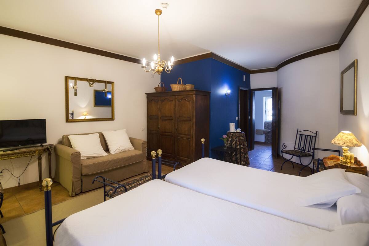 A hotel room with a large bed, a sofa, a television, a wardrobe, and a chandelier.