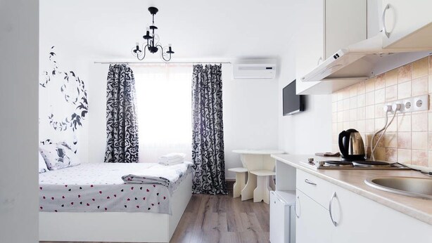 A compact bedroom with a bed, a bedside table, and a window with patterned curtains. Adjacent to the bedroom is a kitchenette with a sink, a stove, and a small countertop. - Odessa Rent Service Apartments at Sea-side (Odesa)