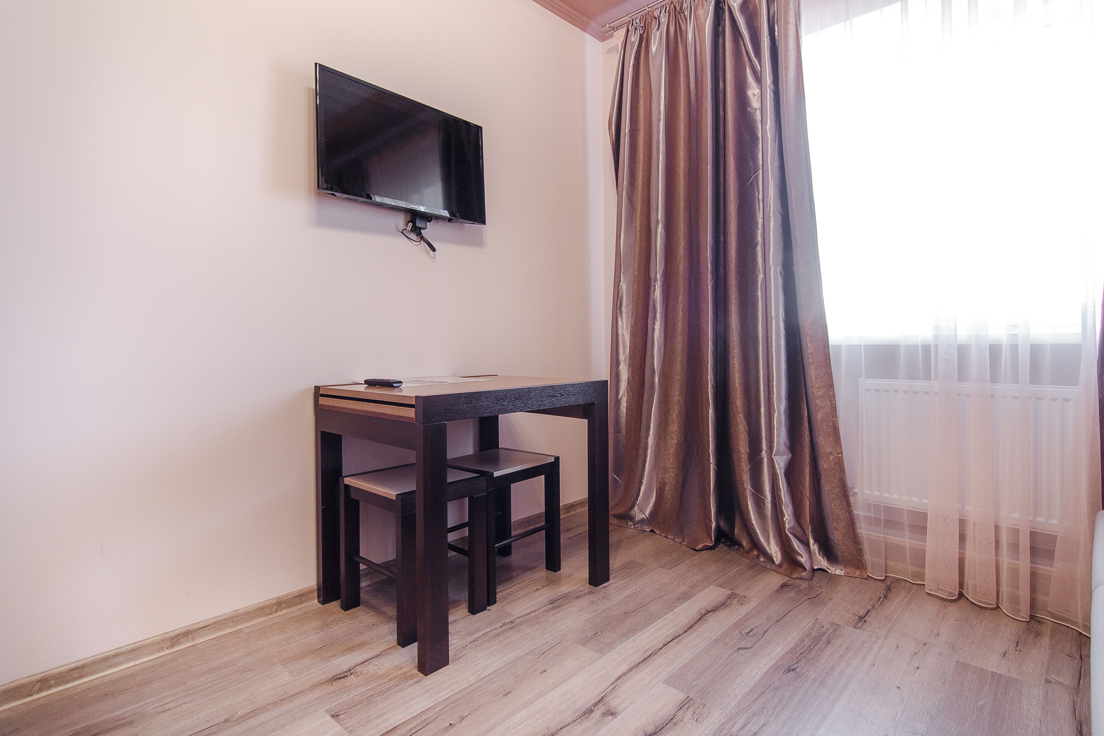 Una habitación con piso de madera, un escritorio de madera oscura con una silla, un televisor de pantalla plana montado en la pared y una ventana con cortinas translúcidas.