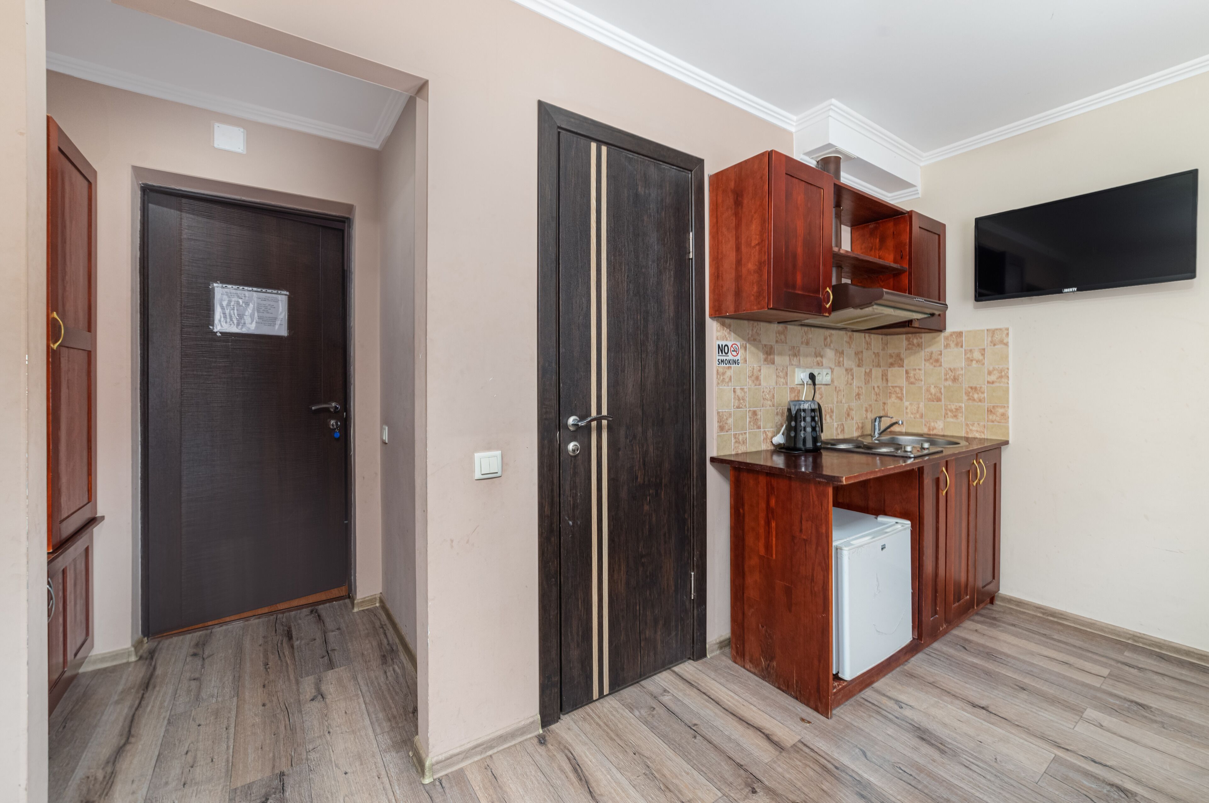 A compact kitchen with wooden cabinets, a built-in refrigerator, and a flat-screen TV mounted above the counter.
