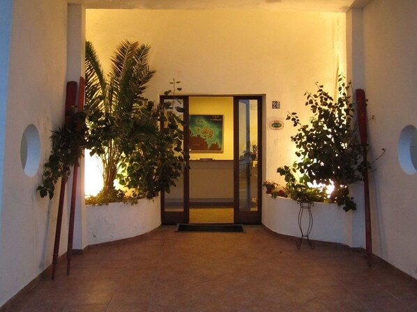 A hallway with a door leading to a room, flanked by plants and a wall-mounted number sign.