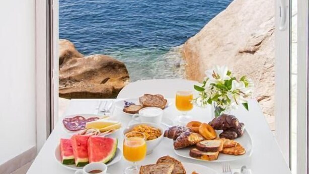 A breakfast table with a variety of foods and a glass of orange juice, set against a backdrop of a rocky coastline and clear blue sea. - Hotel Il Gabbiano (La Maddalena)