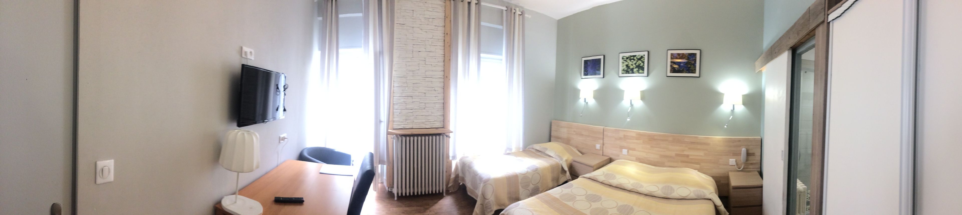 Habitación de hotel con cama, escritorio, silla, radiador, televisor y tres lámparas fijadas a la pared.